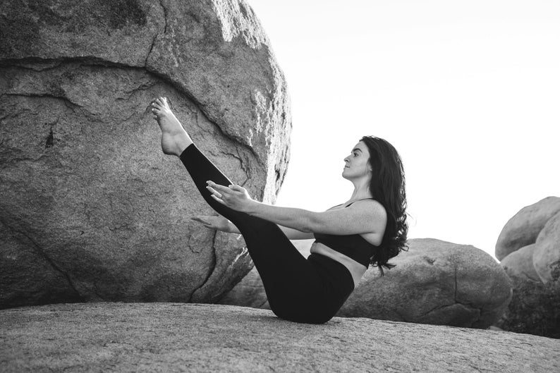 Pu&ograve; includere: Una persona in abbigliamento sportivo nero esegue una posa yoga all'aperto. La persona &egrave; seduta su una roccia, con le gambe estese verso l'alto e le braccia tese. Lo sfondo presenta grandi formazioni rocciose.