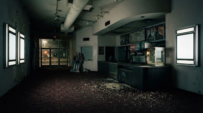 May include: An abandoned cinema lobby featuring a dark carpet with scattered popcorn, a concession stand with a soda fountain and popcorn machine, illuminated wall-mounted display cases, and a glass entrance door visible in the distance.