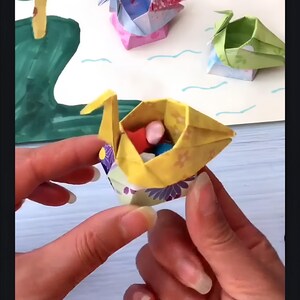 May include: A yellow origami swan with a floral pattern holds small colorful balls inside. The swan is sitting in the palm of a person's hand.