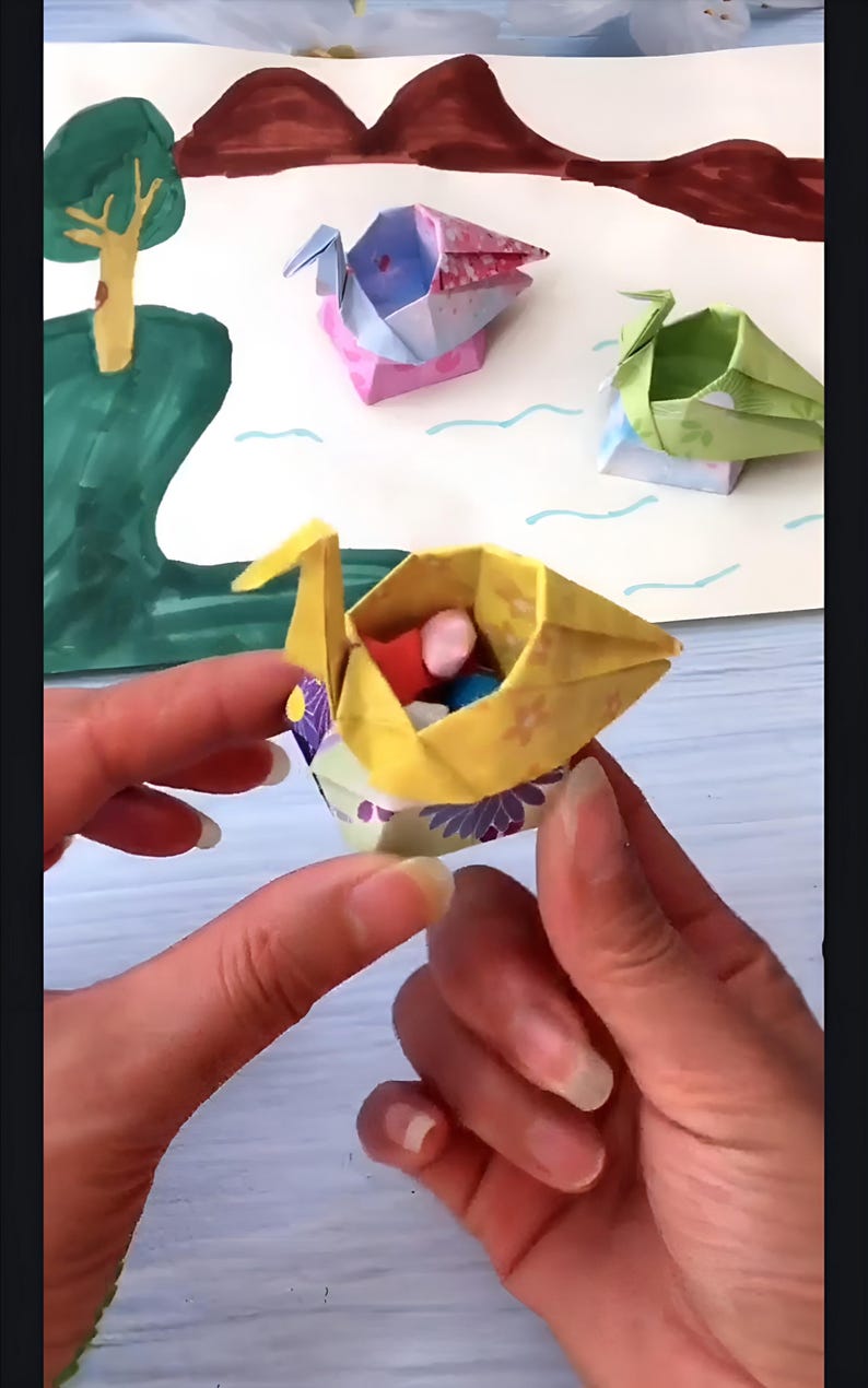 May include: A yellow origami swan with a floral pattern holds small colorful balls inside. The swan is sitting in the palm of a person's hand.