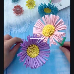 May include: Three paper flowers with yellow centers and pink, purple, and blue petals. The flowers are made from strips of paper and are arranged in a bouquet.