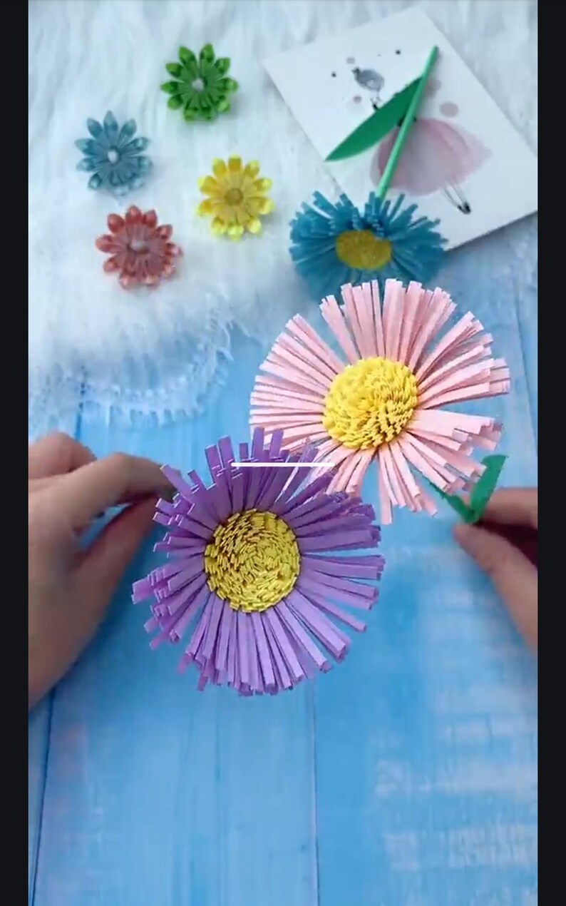 May include: Three paper flowers with yellow centers and pink, purple, and blue petals. The flowers are made from strips of paper and are arranged in a bouquet.