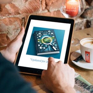 Puede incluir: Una tableta muestra la portada de un libro con un dise&ntilde;o de lupa. El libro es oscuro con detalles blancos y azules. El texto en la pantalla dice "Updated for 2023". Tambi&eacute;n se ven una taza de caf&eacute; y una vela encendida.