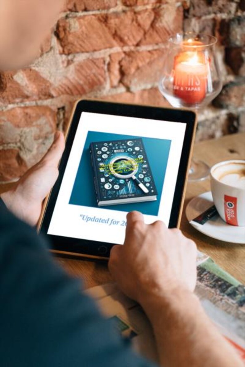 Puede incluir: Una tableta muestra la portada de un libro con un dise&ntilde;o de lupa. El libro es oscuro con detalles blancos y azules. El texto en la pantalla dice "Updated for 2023". Tambi&eacute;n se ven una taza de caf&eacute; y una vela encendida.