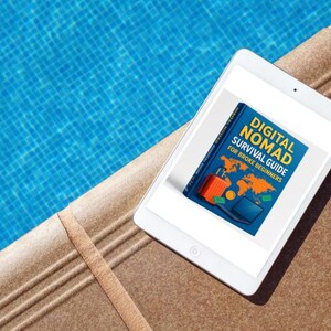 Op de afbeelding: Een witte tablet met de cover van een boek getiteld "DIGITAL NOMAD SURVIVAL GUIDE FOR BROKE BEGINNERS". De cover toont een wereldkaart, een koffer en een laptop. De tablet ligt op een bruin oppervlak naast een blauw zwembad.