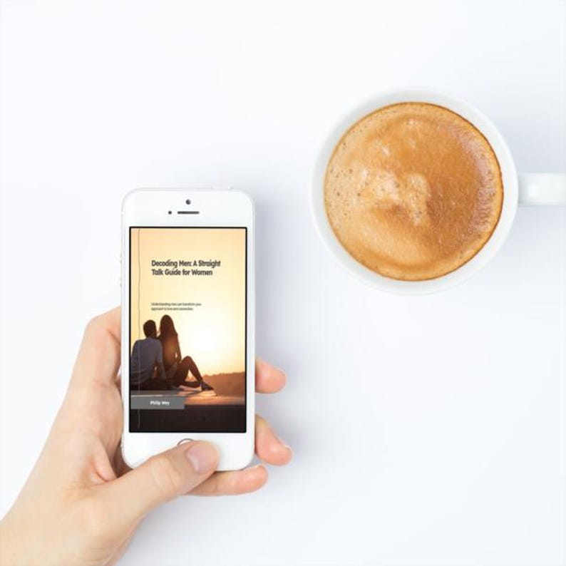 May include: A white smartphone displaying the text "Decoding Men: A Straight Talk Guide for Women" next to a white coffee mug filled with a brown beverage. The phone screen shows a couple silhouetted against a sunset. The background is white.