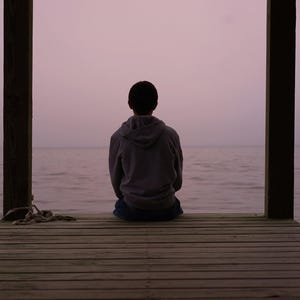 Pu&ograve; includere: Una persona con una felpa grigia con cappuccio &egrave; seduta su un molo di legno, di fronte a una distesa d'acqua calma. Il cielo &egrave; di un morbido viola e la scena &egrave; incorniciata da due pali di legno. Una corda &egrave; appoggiata sul molo.