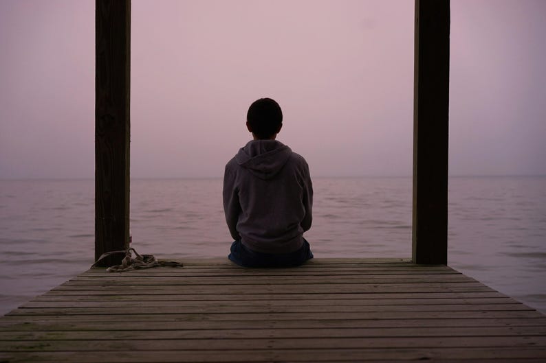 Pu&ograve; includere: Una persona con una felpa grigia con cappuccio &egrave; seduta su un molo di legno, di fronte a una distesa d'acqua calma. Il cielo &egrave; di un morbido viola e la scena &egrave; incorniciata da due pali di legno. Una corda &egrave; appoggiata sul molo.