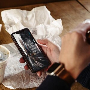 May include: A person holding a smartphone displaying the text "NYC Uncovered: 1001 Bizarre Facts and Hidden Histories." The phone is black with a dark screen. A McDonald's cup with a straw and a burger are also visible.