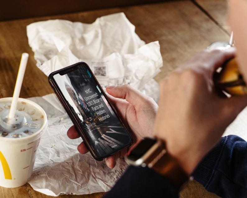 May include: A person holding a smartphone displaying the text "NYC Uncovered: 1001 Bizarre Facts and Hidden Histories." The phone is black with a dark screen. A McDonald's cup with a straw and a burger are also visible.