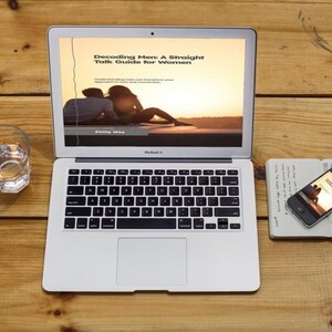 May include: A laptop displaying "Decoding Men: A Straight Talk Guide for Women" on a wooden surface. A glass of water, a smartphone, and a notebook are also on the table. The laptop screen shows a couple at sunset.