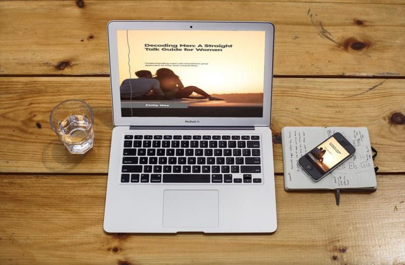 May include: A laptop displaying "Decoding Men: A Straight Talk Guide for Women" on a wooden surface. A glass of water, a smartphone, and a notebook are also on the table. The laptop screen shows a couple at sunset.
