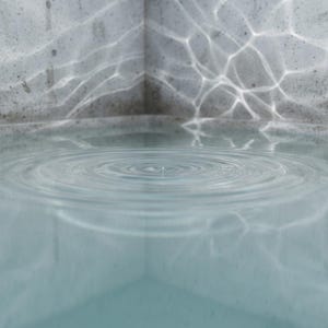 May include: A clear pool of water contained within a grey concrete corner structure. The surface of the water displays concentric circular ripples emanating from a central point, while light reflects off the water to create patterns on the walls.