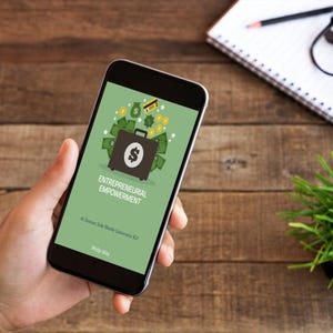 May include: A smartphone screen shows the words "Entrepreneurial Empowerment" and a briefcase graphic with a dollar sign, surrounded by money and a credit card. The phone is held in a hand, with a wooden background.
