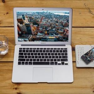 Op de afbeelding: Een zilveren MacBook Air laptop met een stadsbeeld, naast een glas water, een smartphone en een notitieboek op een houten oppervlak. De smartphone en laptop tonen vergelijkbare stadsgezichten. Het notitieboek heeft handgeschreven notities.