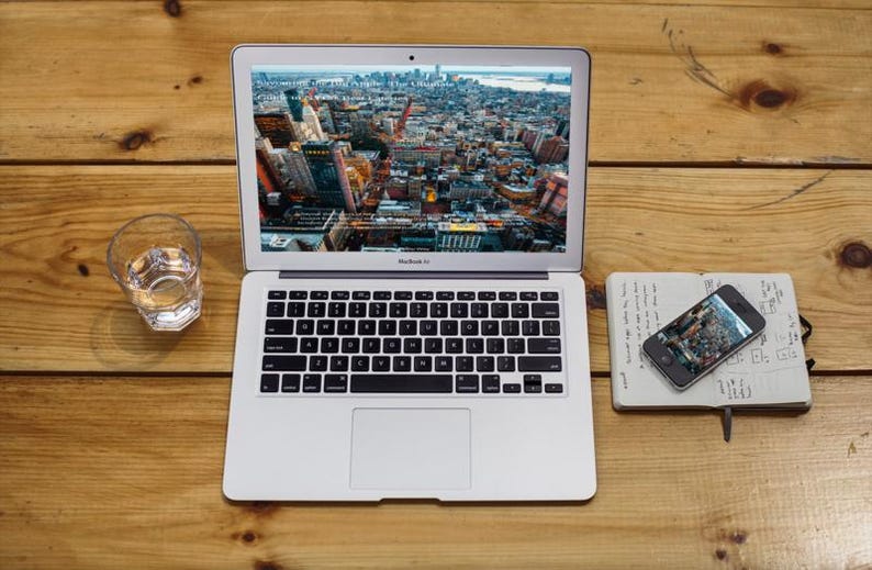 Op de afbeelding: Een zilveren MacBook Air laptop met een stadsbeeld, naast een glas water, een smartphone en een notitieboek op een houten oppervlak. De smartphone en laptop tonen vergelijkbare stadsgezichten. Het notitieboek heeft handgeschreven notities.