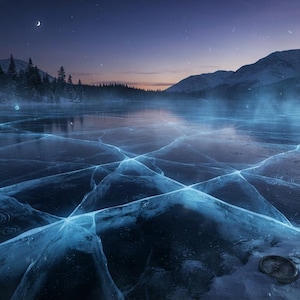 May include: A frozen lake surface featuring deep blue cracks and glowing light patterns under a twilight sky. A small glass lantern and a circular metal object rest on the snowy bank in the foreground, with dark pine trees and mountains in the distance.