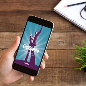以下が含まれることがあります： スマートフォンには、「Confidence」というタイトルの本の表紙が表示されており、スーツを着た人物のグラフィックが描かれています。携帯電話は手に持たれており、背景には木製のテーブル、メモ帳、メガネ、小さな植物があります。