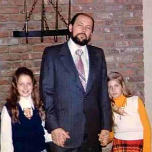May include: A family portrait showing a man in a pinstripe suit, white shirt, and patterned tie, with two young girls. The girls are wearing a white shirt with a navy blue vest and a white shirt with an orange turtleneck and plaid skirt. The background is a brick wall.