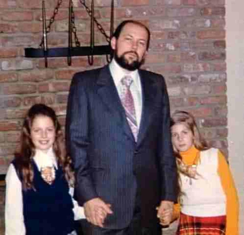 May include: A family portrait showing a man in a pinstripe suit, white shirt, and patterned tie, with two young girls. The girls are wearing a white shirt with a navy blue vest and a white shirt with an orange turtleneck and plaid skirt. The background is a brick wall.