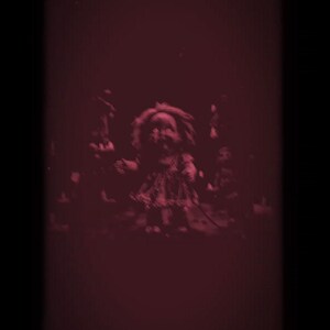 Puede incluir: Imagen oscura y te&ntilde;ida de rojo que presenta una mu&ntilde;eca vintage con una apariencia angustiada. La mu&ntilde;eca es el foco central, rodeada de otras figuras borrosas, creando una est&eacute;tica espeluznante y de terror. La composici&oacute;n general evoca una sensaci&oacute;n de pavor.