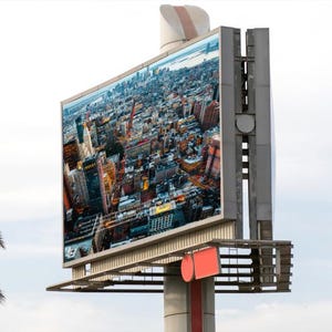 Op de afbeelding: Een groot reclamebord in de openlucht toont een luchtfoto van een stad. De metalen constructie die het bord ondersteunt, heeft rode en witte accenten. Een palmboom is zichtbaar op de voorgrond, tegen een bewolkte lucht.