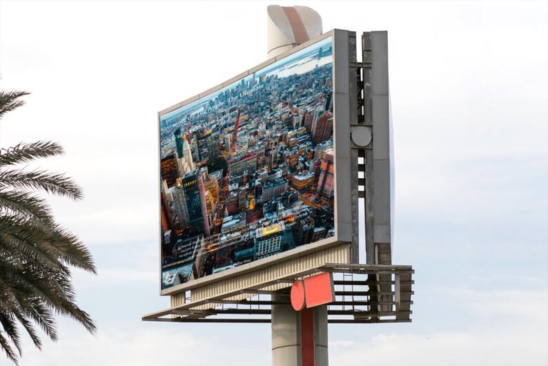 Op de afbeelding: Een groot reclamebord in de openlucht toont een luchtfoto van een stad. De metalen constructie die het bord ondersteunt, heeft rode en witte accenten. Een palmboom is zichtbaar op de voorgrond, tegen een bewolkte lucht.