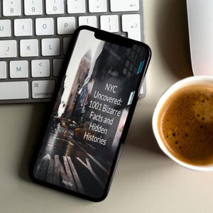 May include: A smartphone displays the cover of a book titled "NYC Uncovered: 1001 Bizarre Facts and Hidden Histories." The phone rests near a white keyboard, a white mouse, and a cup of coffee on a light-colored surface.