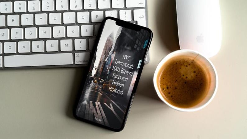 May include: A smartphone displays the cover of a book titled "NYC Uncovered: 1001 Bizarre Facts and Hidden Histories." The phone rests near a white keyboard, a white mouse, and a cup of coffee on a light-colored surface.