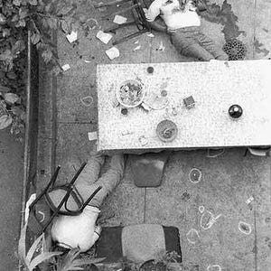 May include: A monochrome image depicting a scene with a person, a table, and scattered objects. A person is slumped in a chair. A table is set with a bowl of food and other items. The ground is covered with debris.