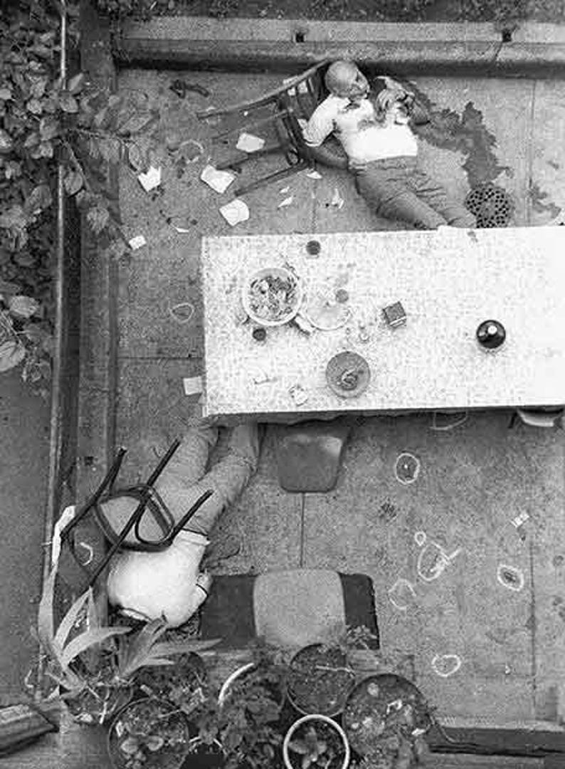 May include: A monochrome image depicting a scene with a person, a table, and scattered objects. A person is slumped in a chair. A table is set with a bowl of food and other items. The ground is covered with debris.