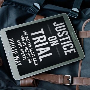 May include: A silver tablet displaying the book cover of "Justice on Trial" by Philip Way. The book cover is black with white text. The tablet rests on a black and brown bag with leather accents.