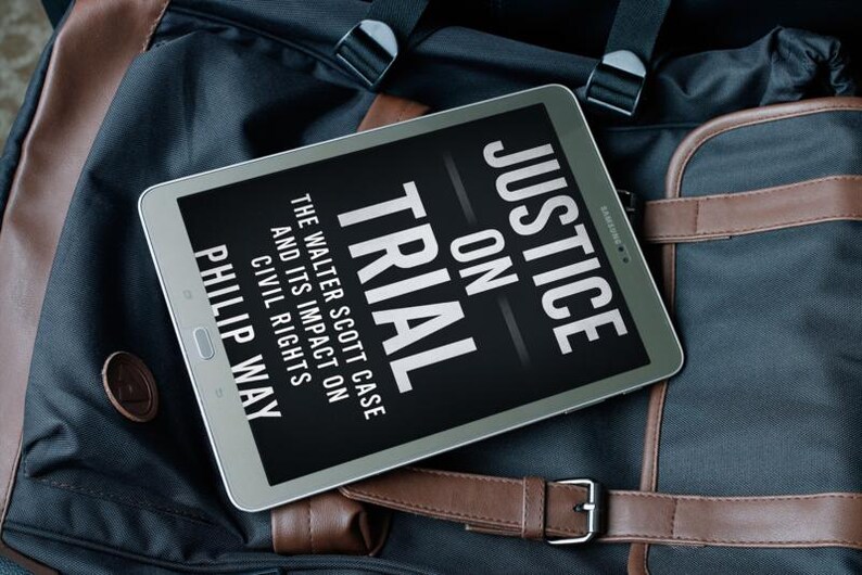 May include: A silver tablet displaying the book cover of "Justice on Trial" by Philip Way. The book cover is black with white text. The tablet rests on a black and brown bag with leather accents.