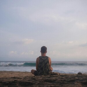 Pu&ograve; includere: Una persona seduta su una spiaggia sabbiosa, rivolta verso l'oceano. La persona indossa una canotta con un motivo mimetico. Il cielo &egrave; nuvoloso e le onde si infrangono in lontananza, creando una scena costiera serena.
