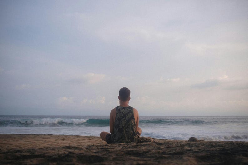 Pu&ograve; includere: Una persona seduta su una spiaggia sabbiosa, rivolta verso l'oceano. La persona indossa una canotta con un motivo mimetico. Il cielo &egrave; nuvoloso e le onde si infrangono in lontananza, creando una scena costiera serena.