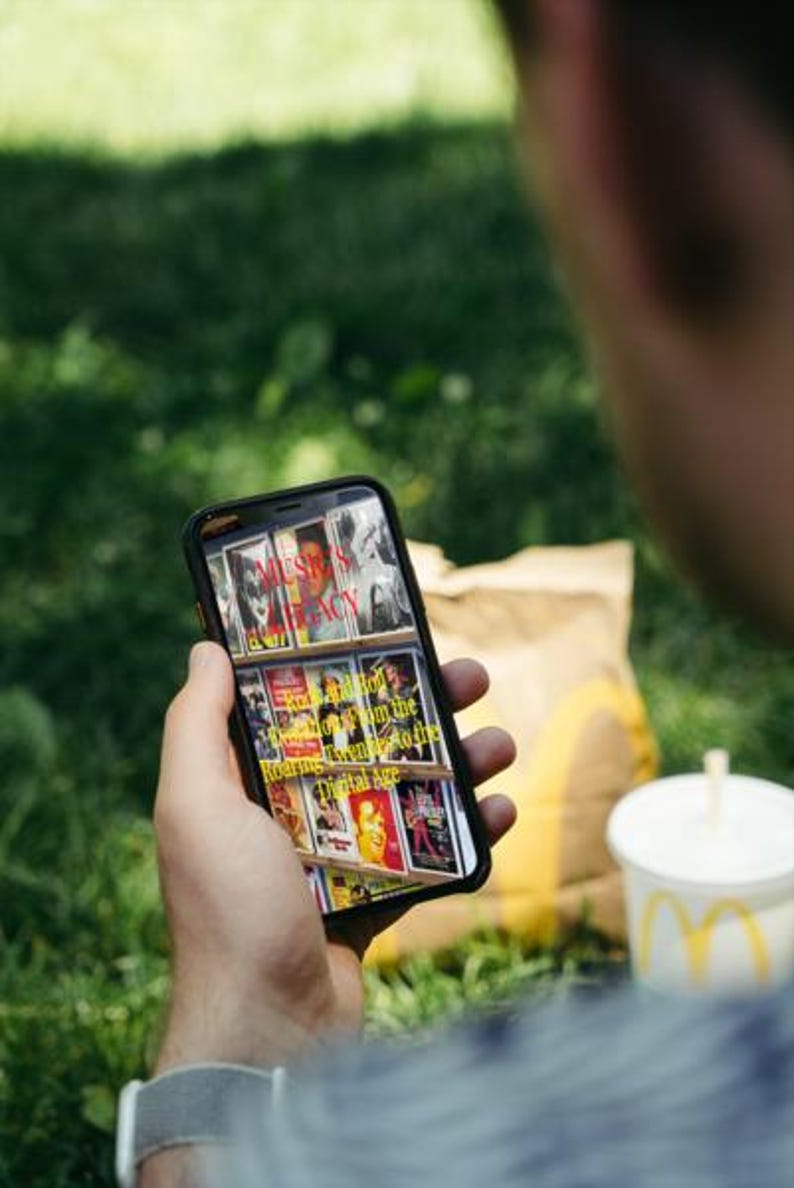 K&ouml;nnte beinhalten: Eine Person h&auml;lt ein Smartphone mit Buchcovern mit dem Text "MUSIC'S LEGACY" und "Rock and Roll from the Roaring Twenties to the Digital Age." Ein McDonald's-Becher und eine T&uuml;te sind im Hintergrund.