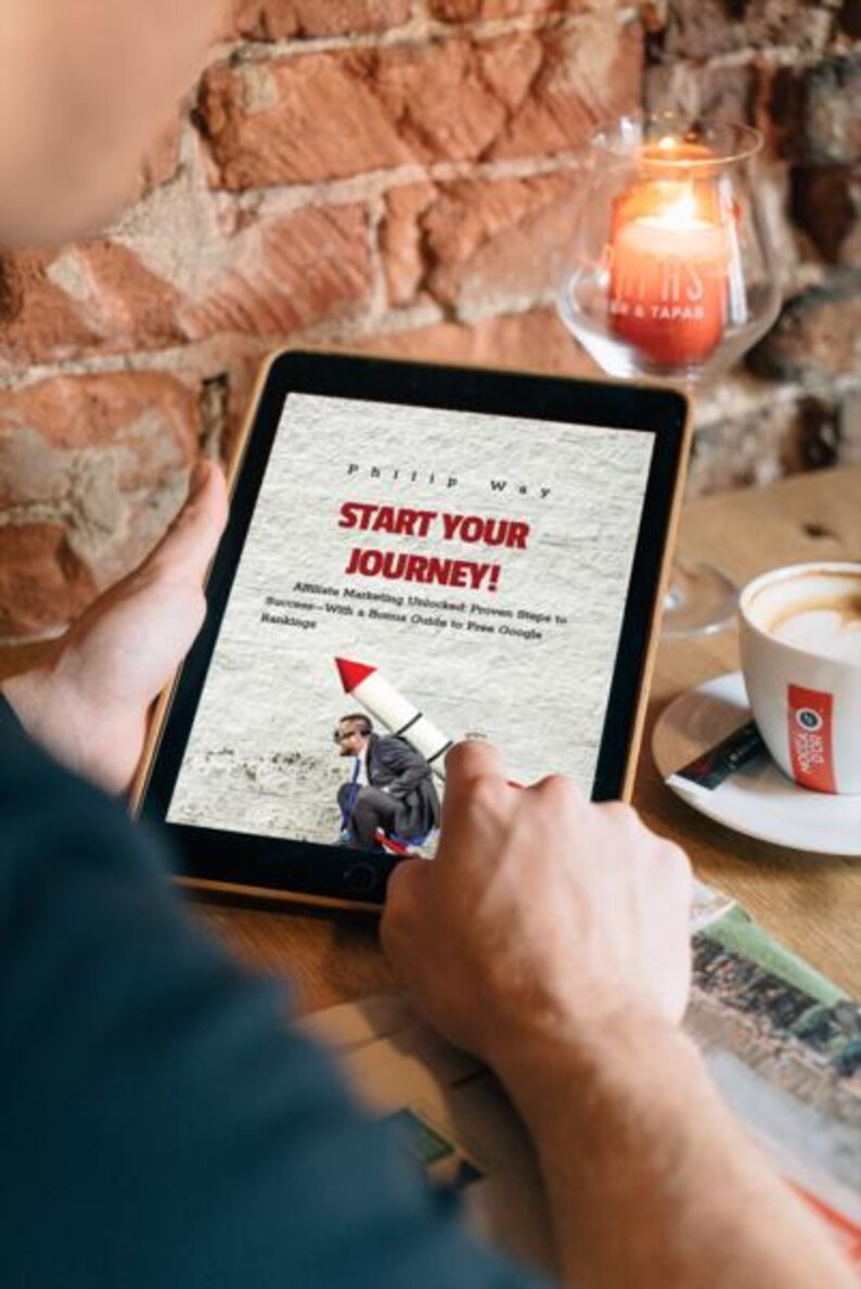 May include: A person holding a tablet displaying the book cover "Start Your Journey!" by Phillip Way. The cover features a man on a rocket. A lit candle and a cup of coffee are in the background.