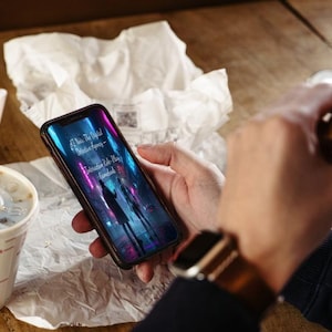 以下が含まれることがあります： ネオンライトで照らされた都市景観を表示するスマートフォン。テキストは「AT Nite: The Official Detective Agency - Interactive Role-Play Gamebook」と表示されています。 電話は、ストローとハンバーガーが入ったマクドナルドのカップの近くに保持されています。