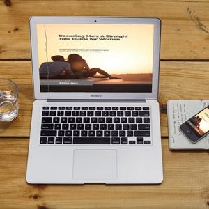 May include: A laptop displaying "Decoding Men: A Straight Talk Guide for Women" on a wooden surface. A glass of water, a smartphone, and a notebook are also on the table. The laptop screen shows a couple at sunset.