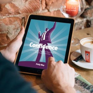 以下が含まれることがあります： フィリップ・ウェイ著「Confidence」の表紙が表示されたタブレット。表紙には、腕を上げたスーツ姿の男性の様式化されたイラストが描かれています。背景には、火の灯ったろうそくとコーヒーカップが見えます。
