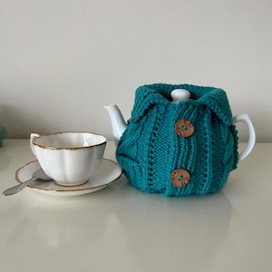 May include: A white ceramic teapot covered by a teal knitted tea cosy featuring a ribbed texture, a folded collar, and two large brown buttons. Beside the teapot sits a white teacup and saucer with a gold rim and a small silver spoon.