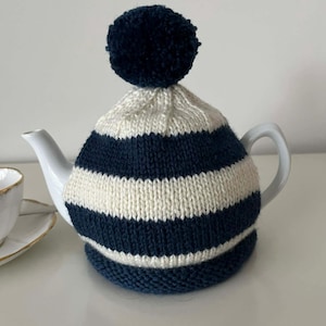 May include: A white ceramic teapot with a navy and cream striped knitted tea cosy. The cosy has a navy pom-pom on top. A white teacup with a gold rim is partially visible in the background.