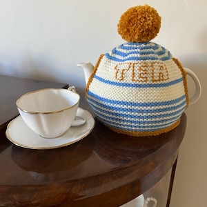 May include: A white porcelain teacup and saucer with gold trim sit beside a teapot with a knitted tea cosy. The cosy is white with blue stripes and the word "TEA" in gold. A large orange pom-pom tops the cosy.