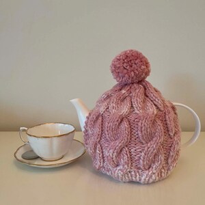May include: A white porcelain teapot with a pink knitted tea cosy and a matching teacup and saucer. The cosy has a cable knit design and a pom-pom on top. The teacup and saucer have gold trim.
