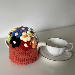 May include: An orange knitted tea cosy decorated with colourful crocheted flowers. A white teacup and saucer with gold edging are placed next to the teapot. The cosy is designed to keep tea warm.