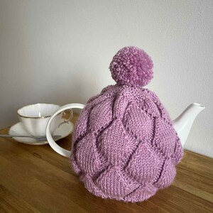 May include: A lilac knitted tea cosy featuring a diamond pattern and a matching pom-pom. The cosy is placed on a white teapot, with a teacup and saucer in the background. The cosy is designed to keep tea warm, a traditional kitchen item.