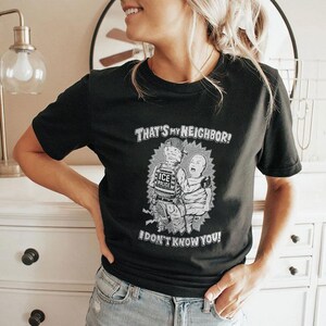 Op de afbeelding: Zwart t-shirt met een grafisch ontwerp met twee cartoonfiguren en de tekst "THAT'S MY NEIGHBOR! I DON'T KNOW YOU!". Het ontwerp is in wit- en grijstinten.