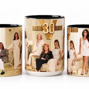 May include: A ceramic mug with a black handle and rim, featuring a wrap-around photographic print of the television show The View. The design includes the gold text THE VIEW and images of the cast members in a bright, indoor setting with furniture.