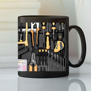 Puede incluir: Taza de cerámica negra con un diseño de caja de herramientas que muestra varias herramientas en amarillo y negro. Las herramientas incluyen una sierra, alicates, destornilladores y una cinta métrica. La taza tiene un asa negra y un acabado brillante.