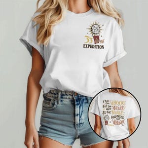 Puede incluir: Camiseta blanca con un gráfico en la parte delantera que dice "33 EXPEDITION" con un diseño de sol y luna. La parte posterior de la camiseta tiene el texto "I TOO AM WHOOO, BUT I'M THERE ALSO SO THE WHEER BALANCES THE WHOOO. ESOME."